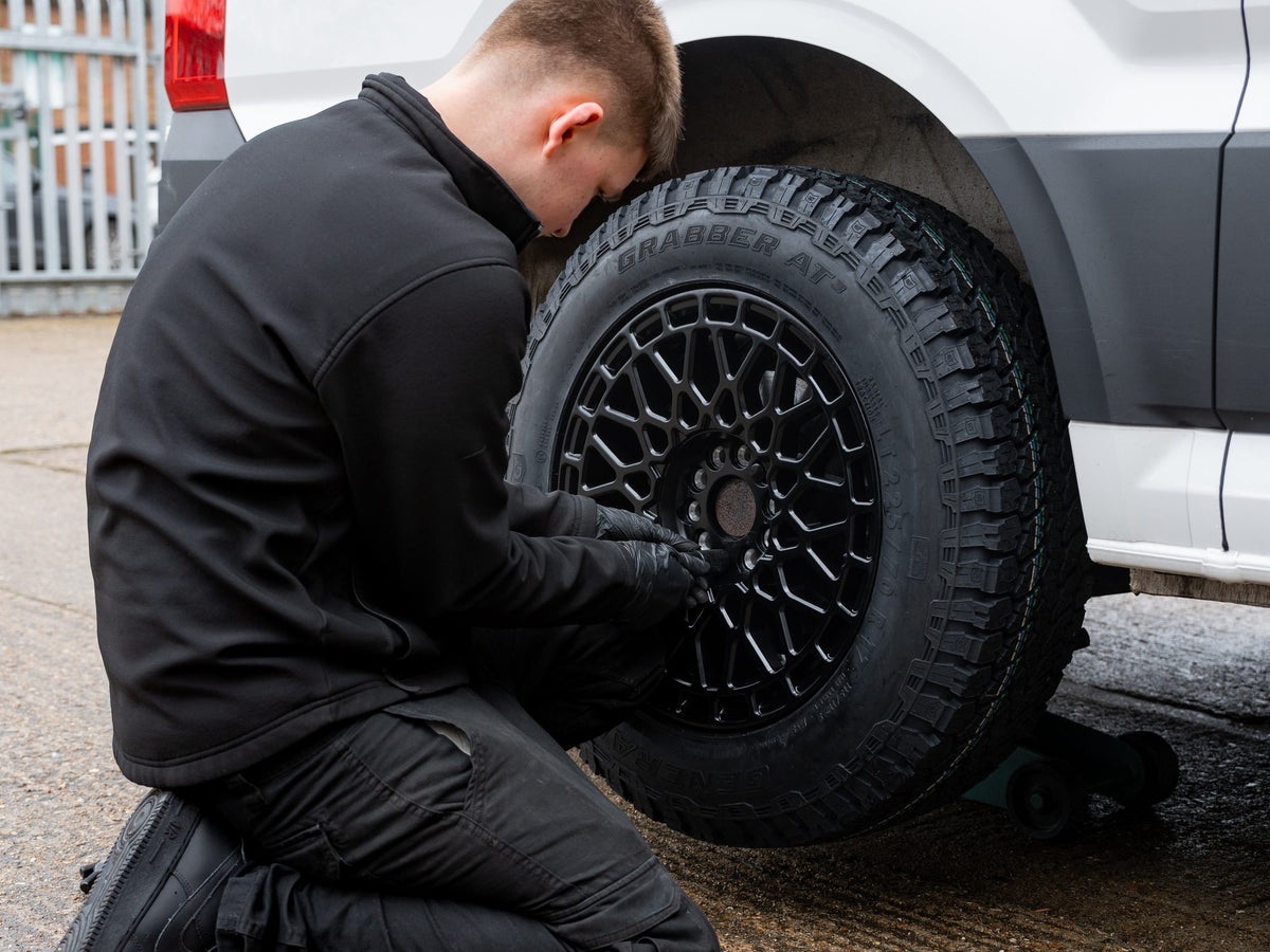 A Rogue fitter kneeling at the arch, fitting a black mesh wheel on a General Grabber AT3 to a white van