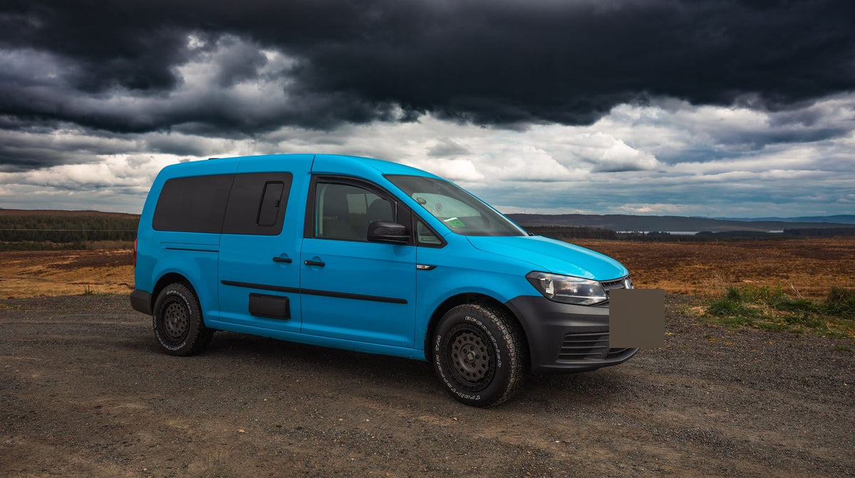 A blue Volkswagen Caddy on Rogue wheels on a gravel road under a dark storm sky