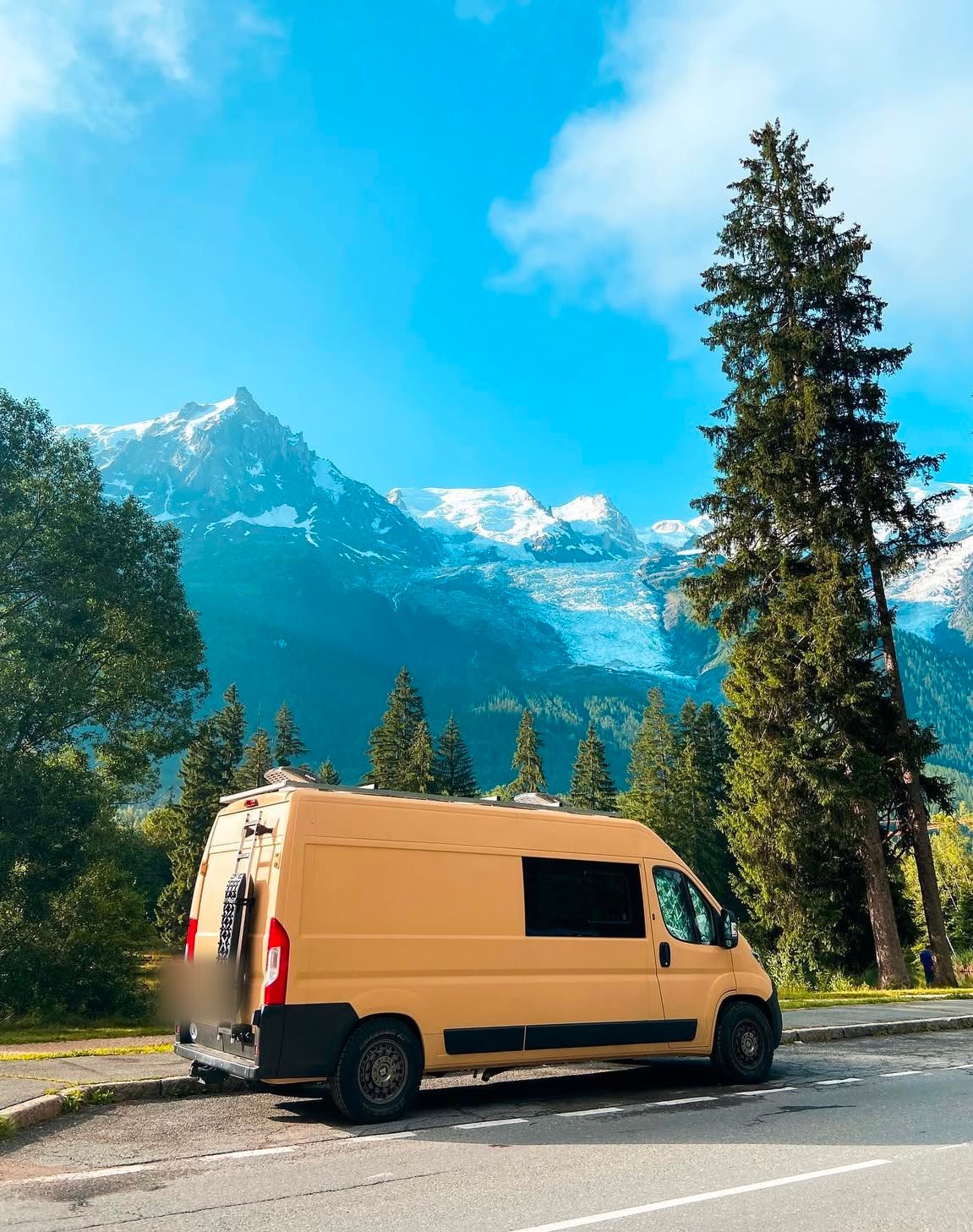 A tan camper van in a lay-by with snow-capped alpine peaks behind