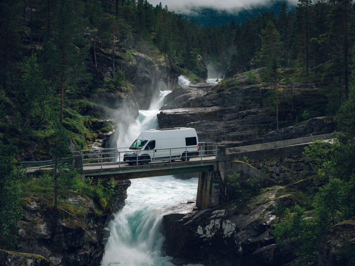 A white camper van crossing a bridge over a waterfall in a forested gorge