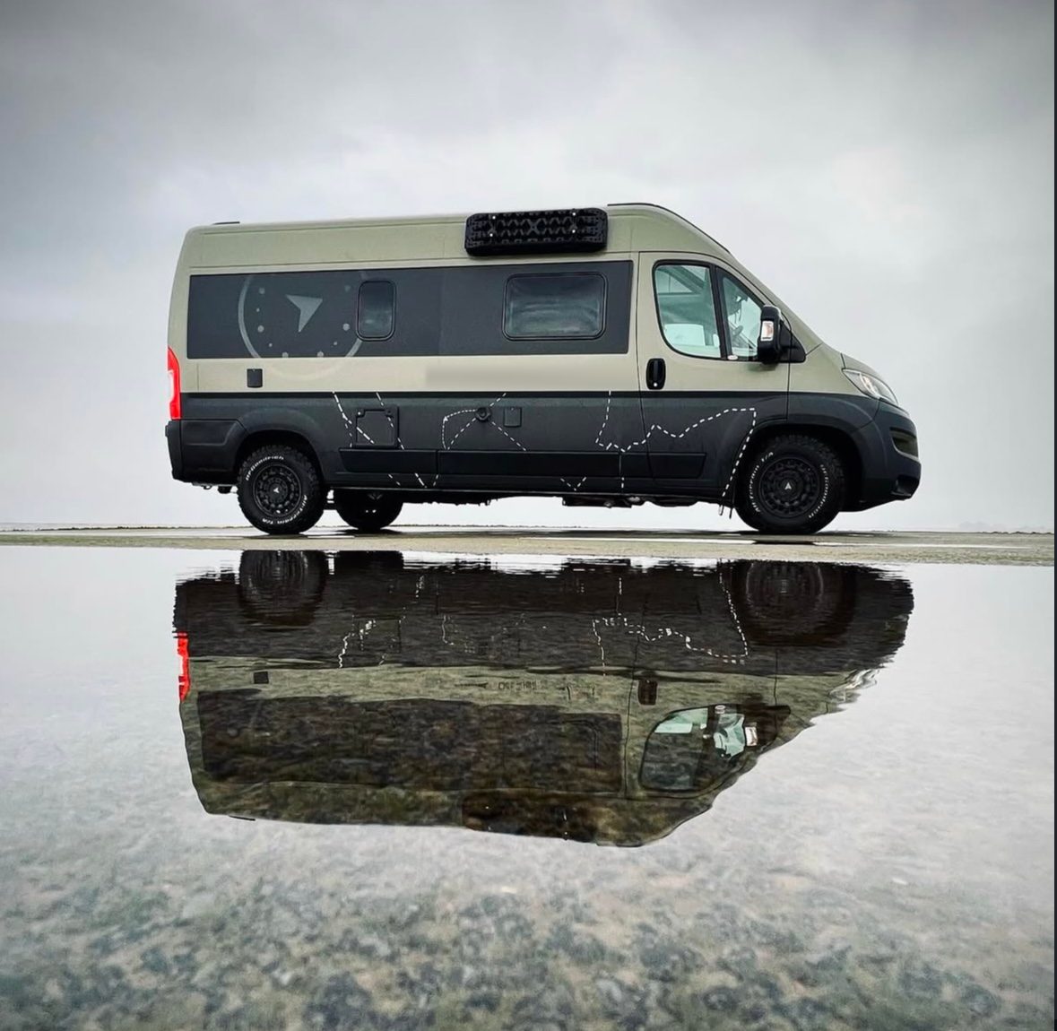 A camper van side-on, reflected in a still lake under cloudy skies