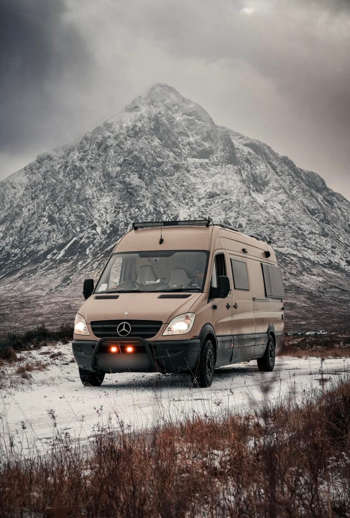A Sprinter camper with a light bar on a snowy track below a rocky peak