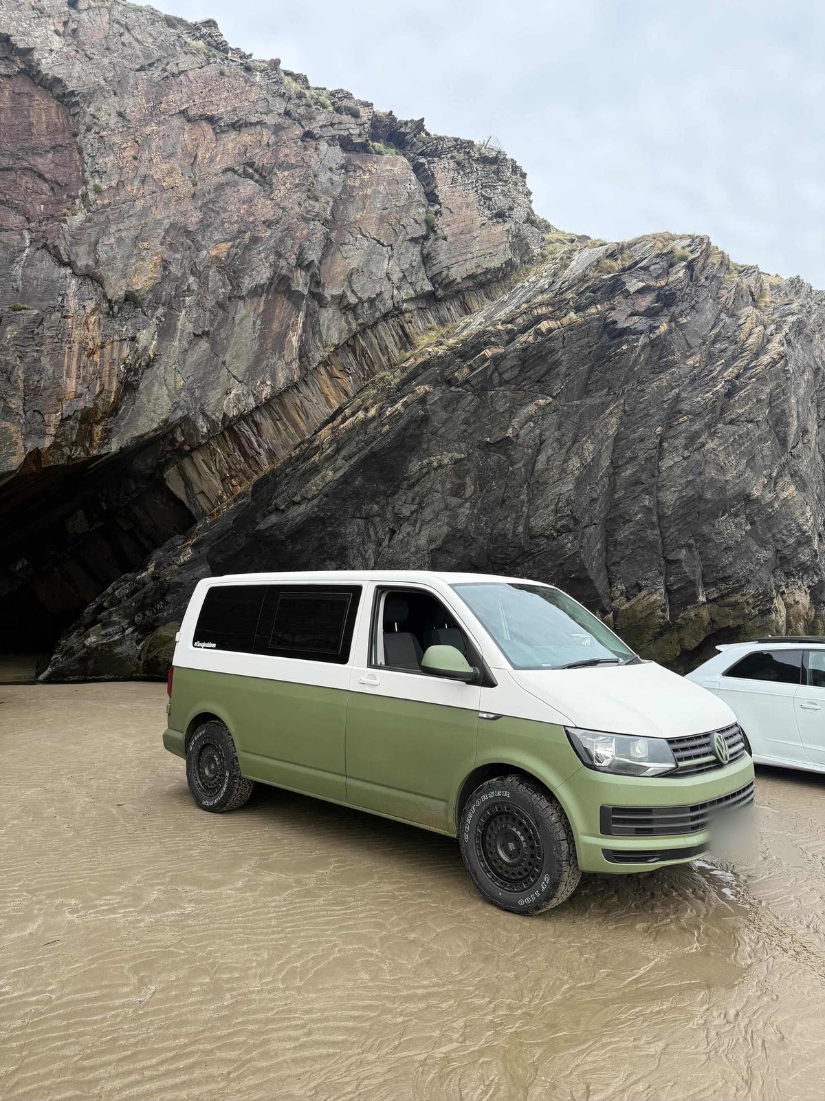 An olive green Volkswagen Transporter on wet sand beside a rocky cliff