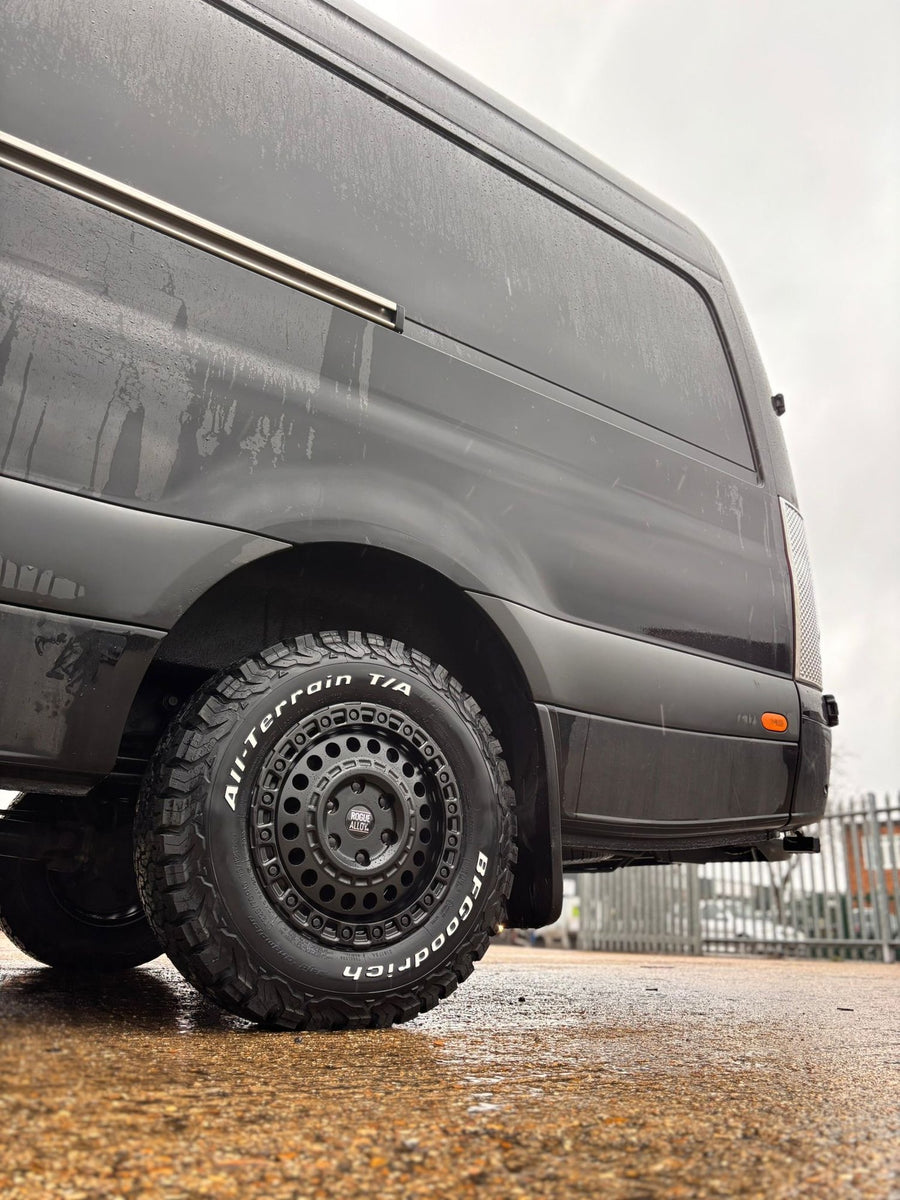 A matte black beadlock-style Rogue wheel on a Sprinter or Crafter-type panel van conversion, BFGoodrich All-Terrain, in a rainy industrial yard
